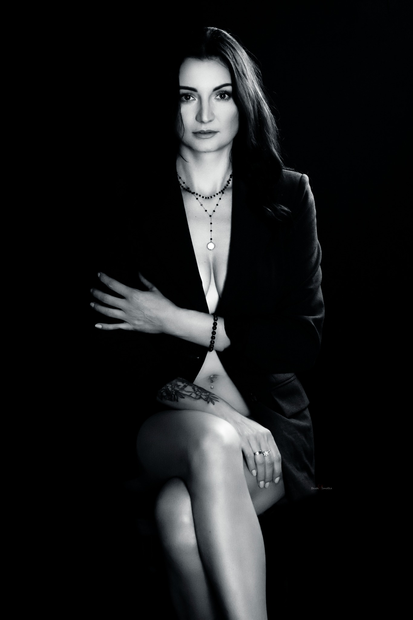 Portret kobiety w czarno-białej tonacji, ubrana w marynarkę, z delikatną biżuterią i tatuażem na ramieniu, na ciemnym tle. Subtelne oświetlenie podkreśla rysy twarzy.