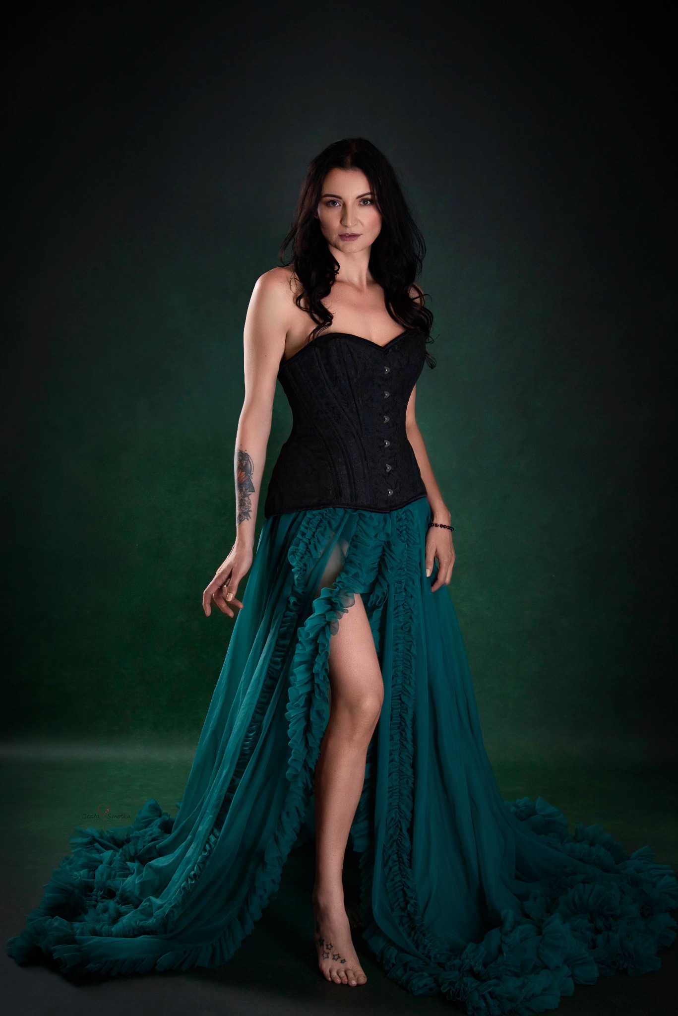 Kobieta w czarnym gorsecie i szmaragdowej, zwiewnej sukni na ciemnozielonym tle. Widoczny tatuaż na ramieniu i stopie. Delikatne światło podkreśla urodę modelki.