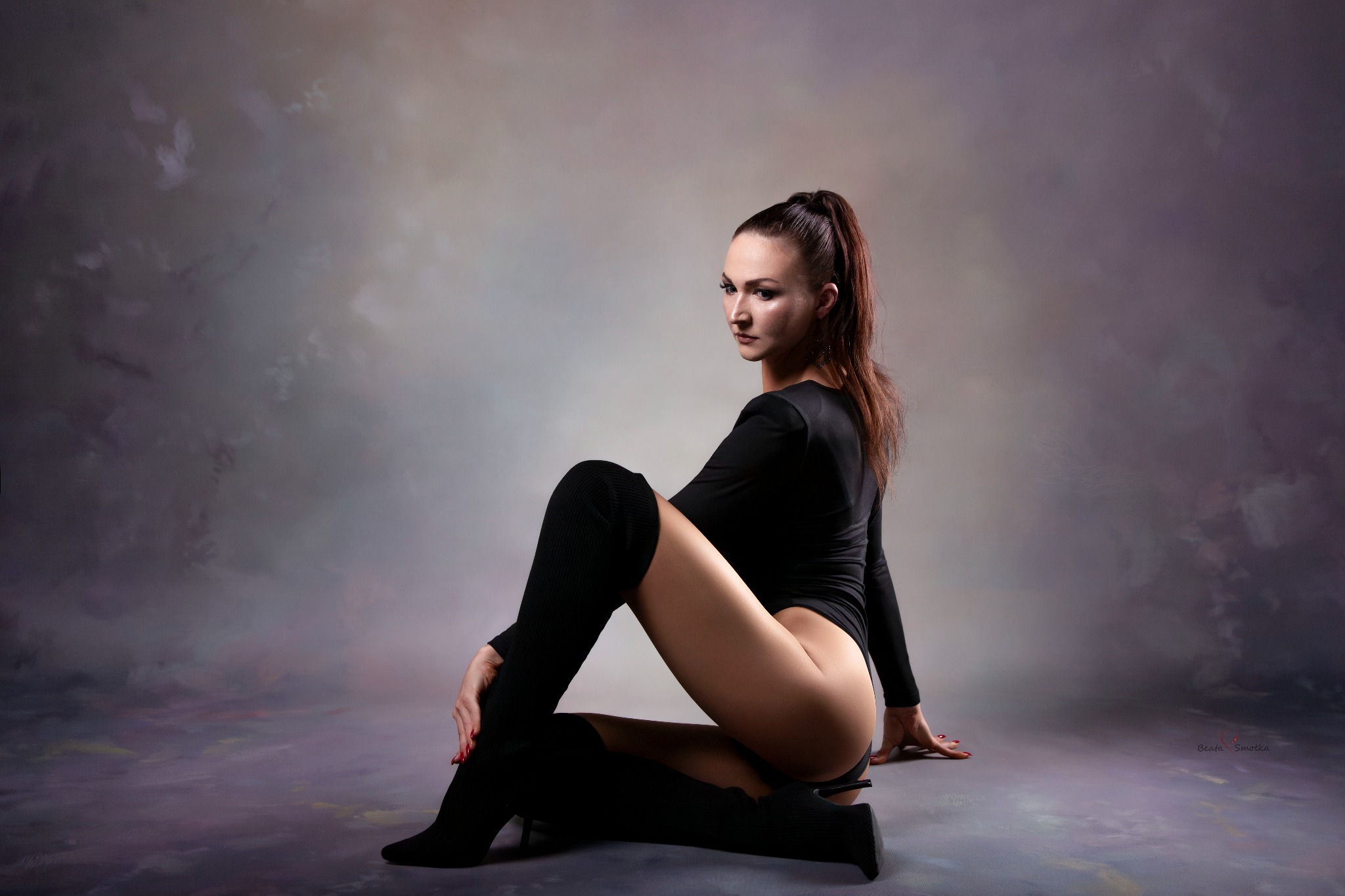 Kobieta w czarnym body i kozakach za kolano siedzi na tle abstrakcyjnego tła, spoglądając przez ramię. Uczesana w wysoki kucyk, z delikatnym makijażem.