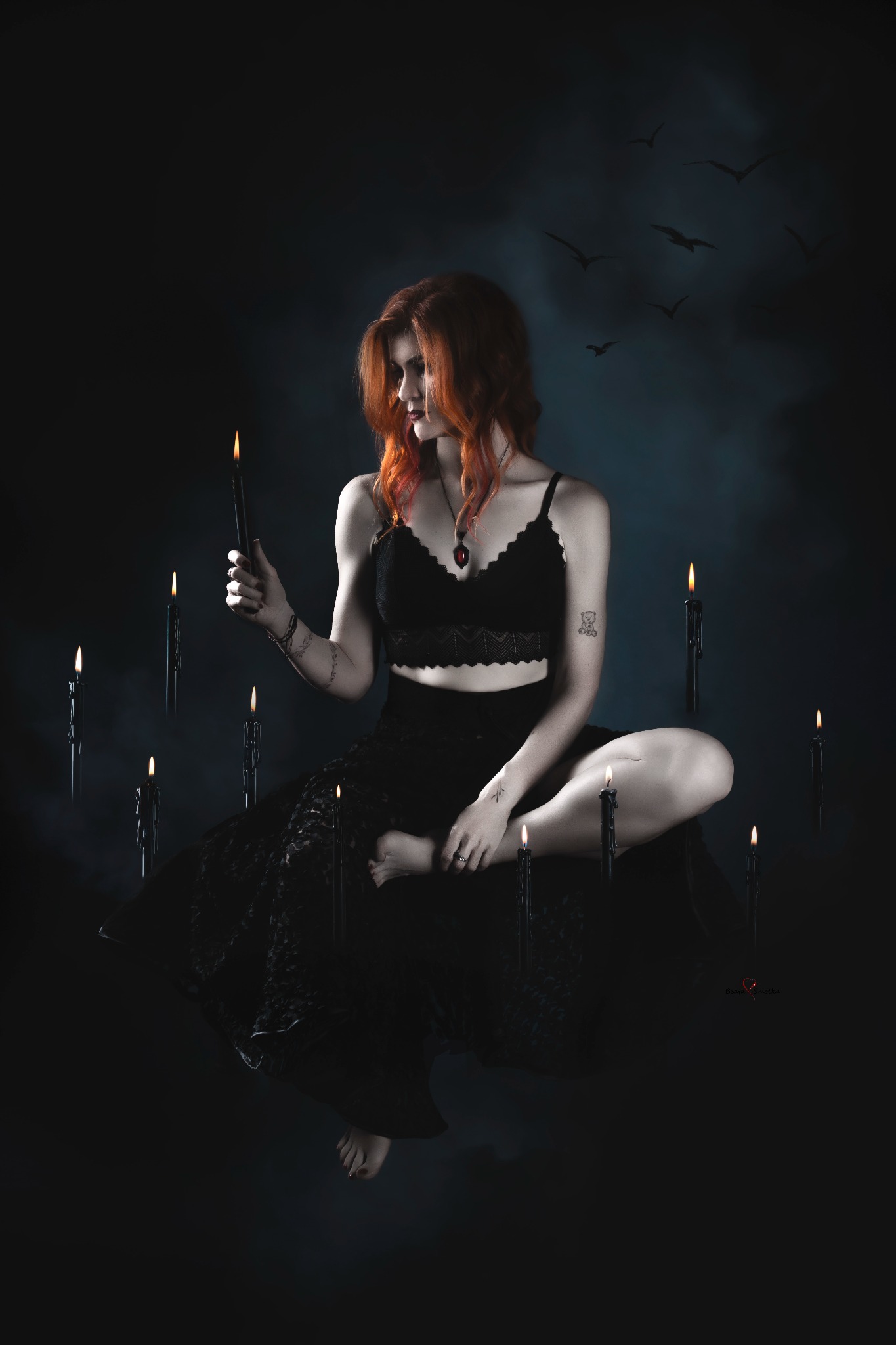 Kobieta z rudymi włosami w czarnym stroju, otoczona płonącymi czarnymi świecami na ciemnym tle z sylwetkami ptaków, tworząca mistyczny klimat.