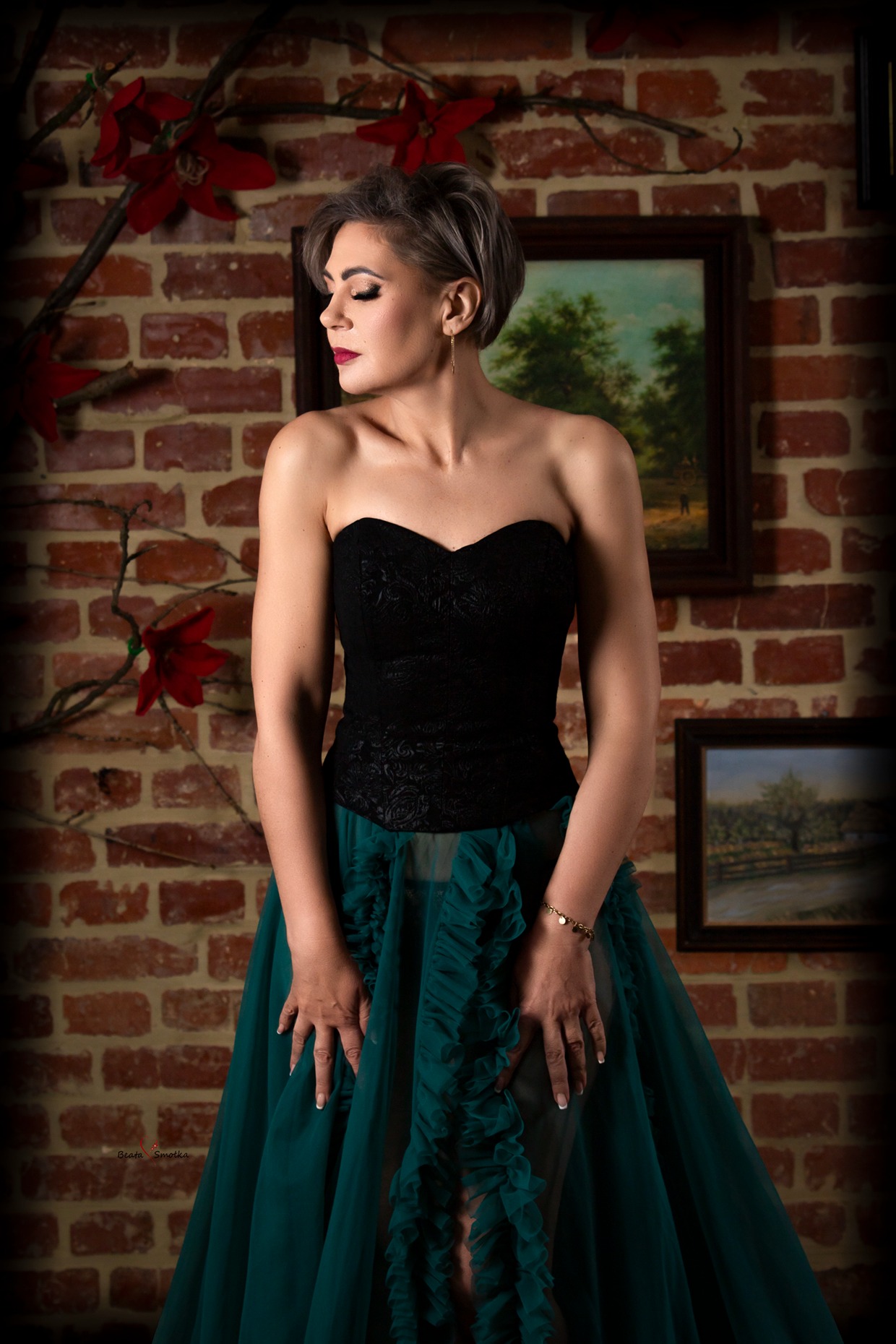 Elegancka kobieta w czarno-zielonej sukni pozuje na tle ceglanej ściany z obrazami, prezentując subtelny makijaż i biżuterię. Stonowane oświetlenie podkreśla detale.