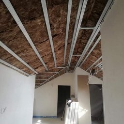 Złota rączka - Stelaż metalowy pod izolację poddasza z wełny mineralnej, widok z dołu. W tle surowe ściany i drzwi, przygotowane do dalszych prac wykończeniowych.