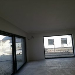 Złota rączka - Surowe wnętrze z dużymi oknami i betonową podłogą, ściany przygotowane do dalszych prac wykończeniowych, widok na zewnątrz przez okna.