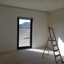 Złota rączka - Pokój po zagruntowaniu ścian, z oknem i drabiną malarską; widoczne taśmy malarskie przy suficie, przygotowanie do dalszych prac.