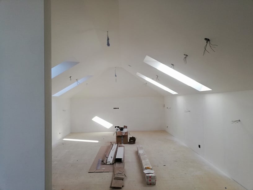 Wnętrze poddasza ze skośnym sufitem i oknami dachowymi, w trakcie prac. Widoczne kable, kartony i materiały budowlane. Świeżo pomalowane ściany na biało.