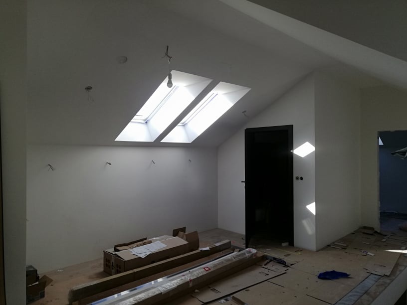 Wnętrze poddasza ze skośnym sufitem i dwoma oknami dachowymi. Widoczne przygotowania do prac wykończeniowych: kartony, profile, zabezpieczona podłoga, czarne drzwi.