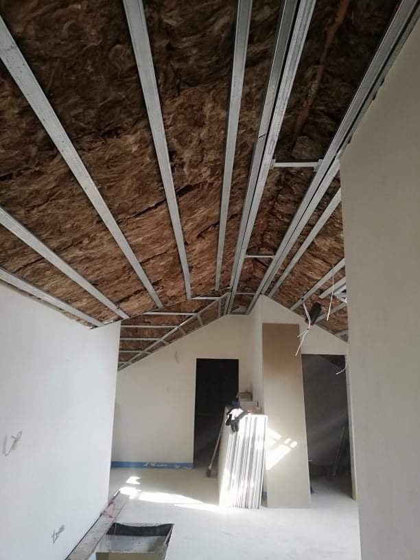 Stelaż metalowy pod izolację poddasza z wełny mineralnej, widok z dołu. W tle surowe ściany i drzwi, przygotowane do dalszych prac wykończeniowych.