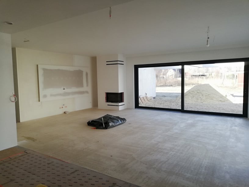 Wnętrze w trakcie wykańczania: ściany po szpachlowaniu, przygotowane podłoże pod posadzkę, duże okno przesuwne z widokiem na zewnątrz, kominek z czarnymi akcentami.