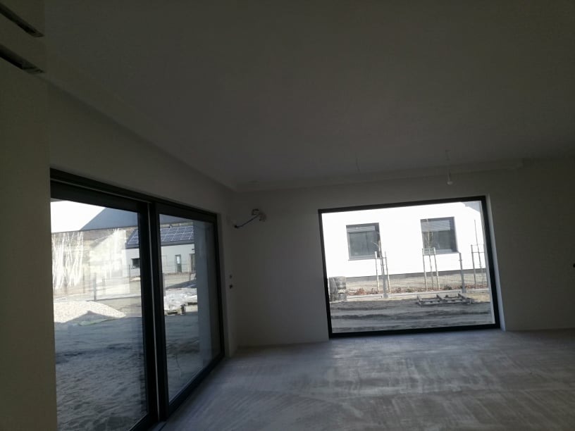 Surowe wnętrze z dużymi oknami i betonową podłogą, ściany przygotowane do dalszych prac wykończeniowych, widok na zewnątrz przez okna.