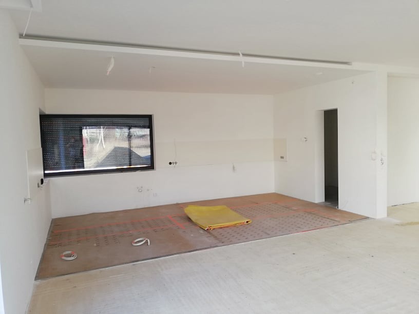 Wnętrze w trakcie remontu: widoczne przewody ogrzewania podłogowego na płycie, surowe ściany z instalacją elektryczną, okno z żaluzjami. Etap prac przed wykończeniem.