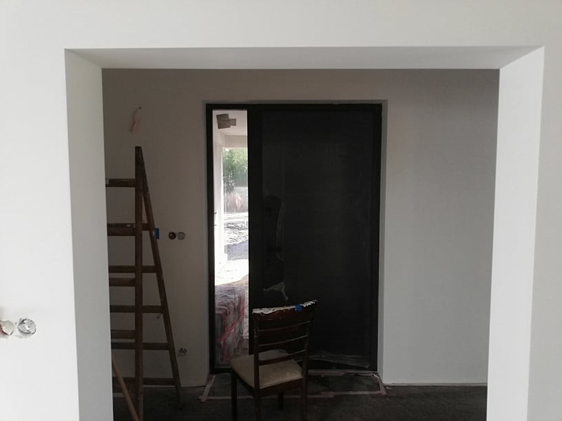 Wnętrze w trakcie prac: ciemne drzwi, ściany pomalowane na jasny kolor, drewniana drabina oparta o ścianę, krzesło przy drzwiach. Widoczne gniazdka elektryczne.