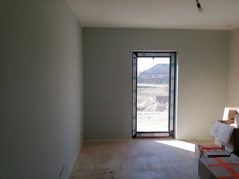Jasne wnętrze z oknem i świeżo pomalowanymi ścianami w odcieniach szarości. Na podłodze widoczne kartony, a na zewnątrz okna krajobraz z zabudowaniami.