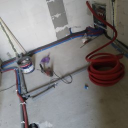 Leszek Kasior - Wnętrze w trakcie instalacji ogrzewania podłogowego: czerwone rury ułożone na szarym betonie, widoczne narzędzia i instalacje w ścianach z białych bloczków.