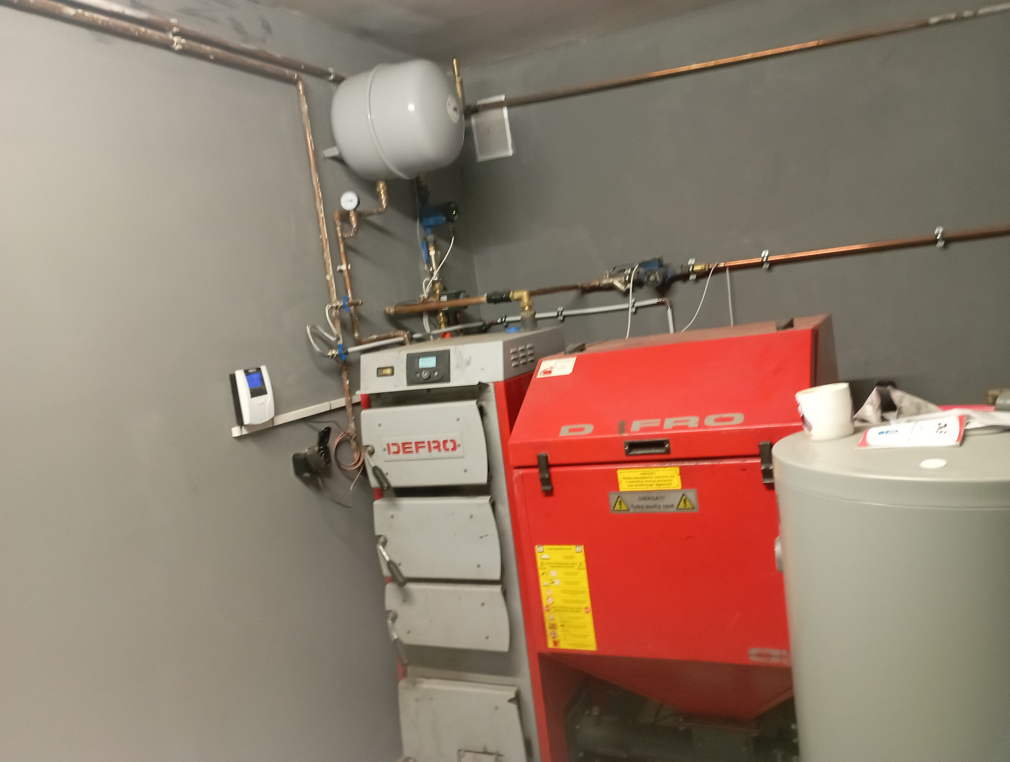 Zmontowany piec CO marki DEFRO z czerwonym podajnikiem i szarym zasobnikiem, widoczne rury instalacji. Szare ściany pomieszczenia.