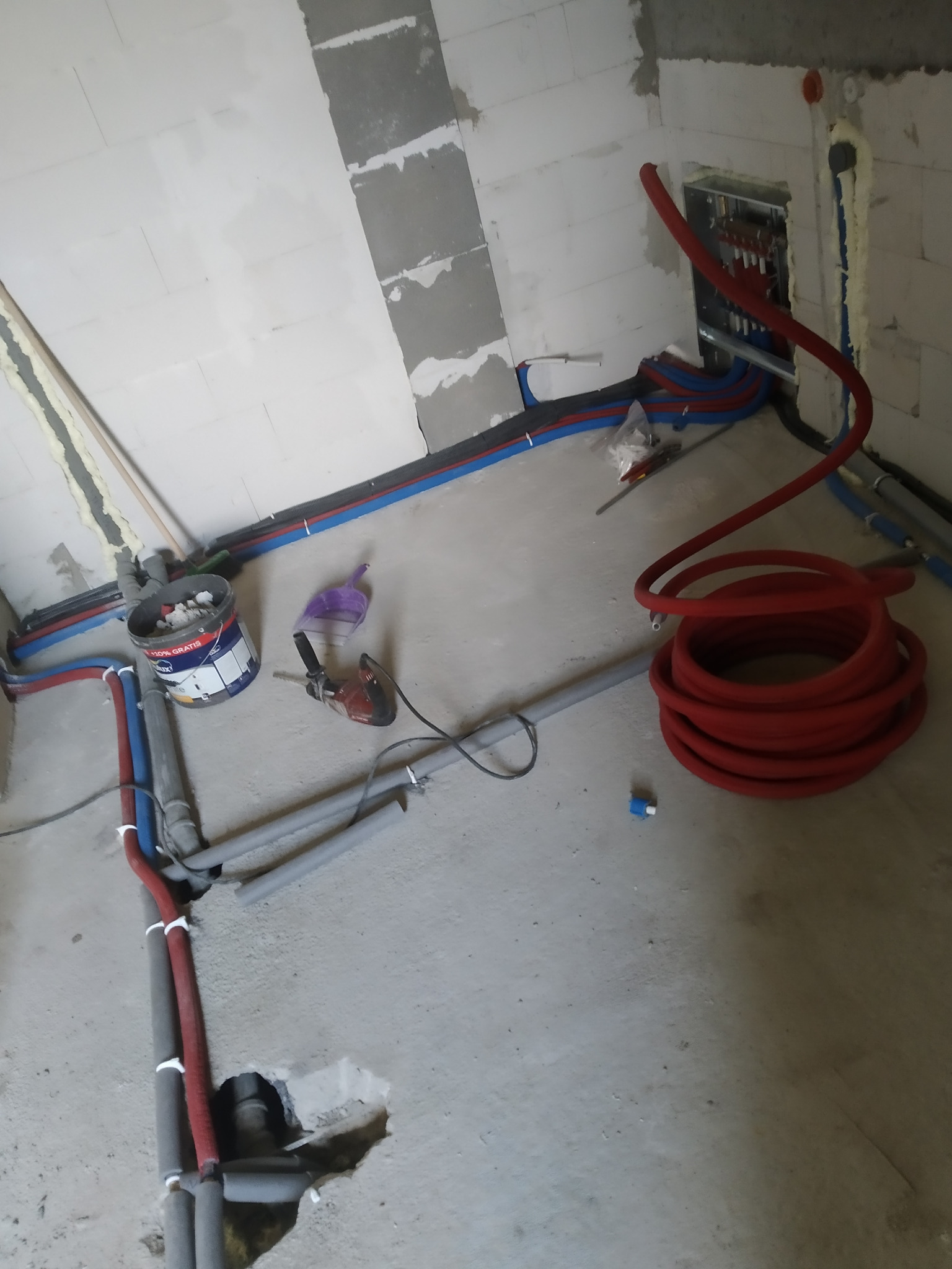 Wnętrze w trakcie instalacji ogrzewania podłogowego: czerwone rury ułożone na szarym betonie, widoczne narzędzia i instalacje w ścianach z białych bloczków.