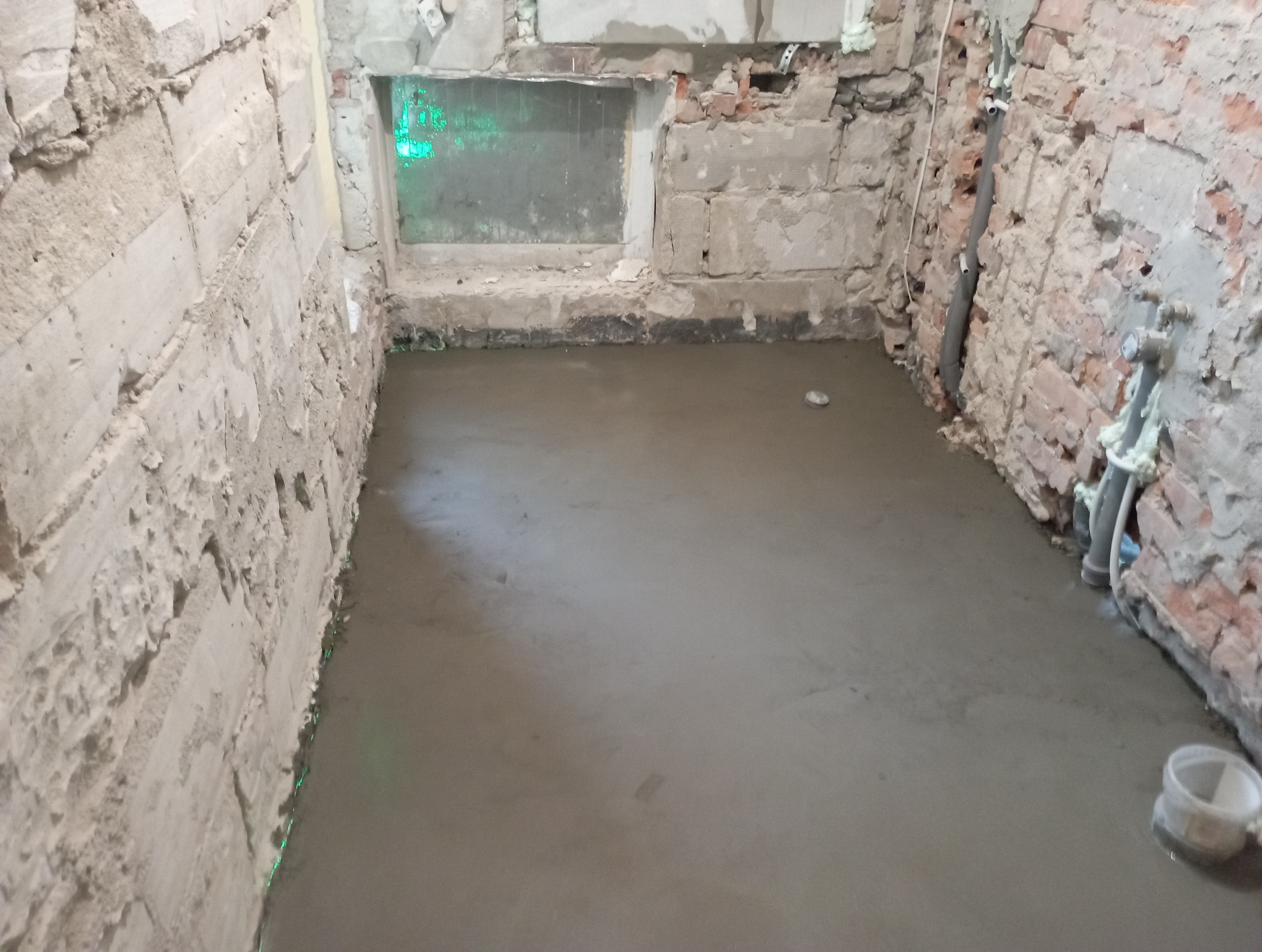 Wyrównana, świeża wylewka na podłodze w pomieszczeniu z odsłoniętymi cegłami i instalacjami. Widoczne rury i niedokończone ściany.