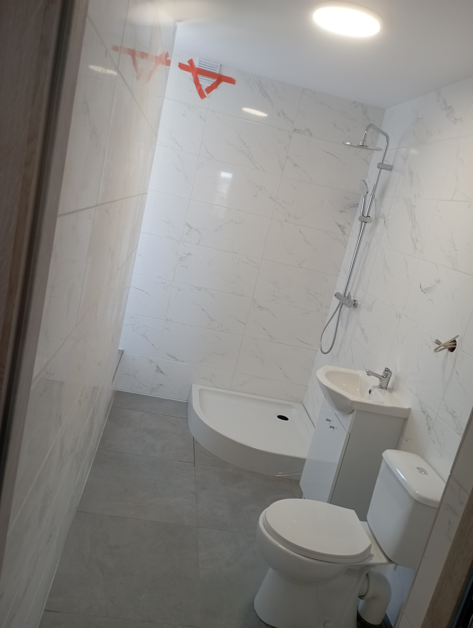 Nowoczesna, jasna łazienka po remoncie: kabina prysznicowa, umywalka z szafką i toaleta. Białe płytki imitujące marmur, szare płytki podłogowe. Widoczna taśma zabezpieczająca.