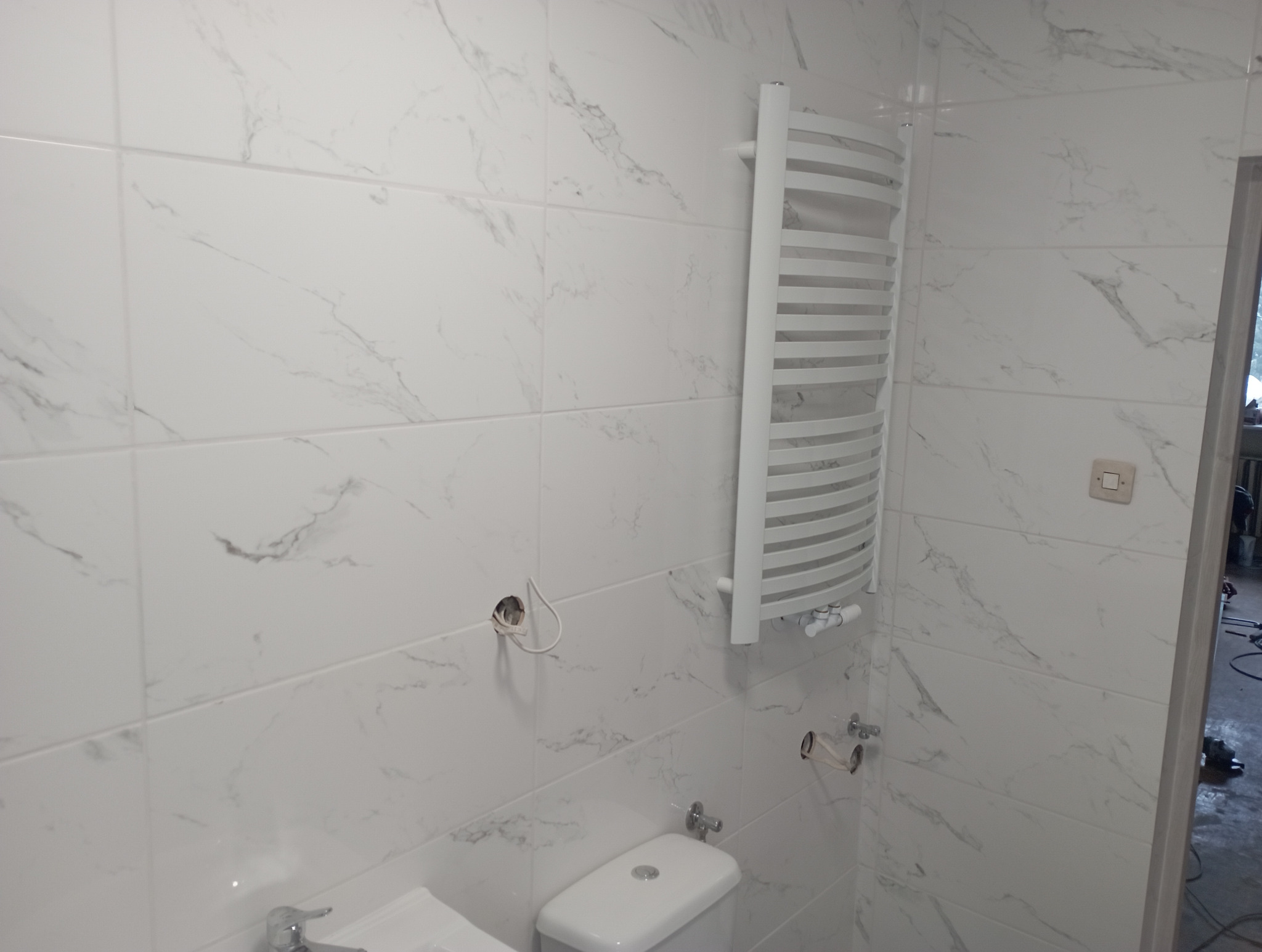 Biała łazienka z grzejnikiem drabinkowym, płytkami imitującymi marmur i przygotowanymi przyłączami hydraulicznymi. Widoczne elementy instalacji przed zabudową.