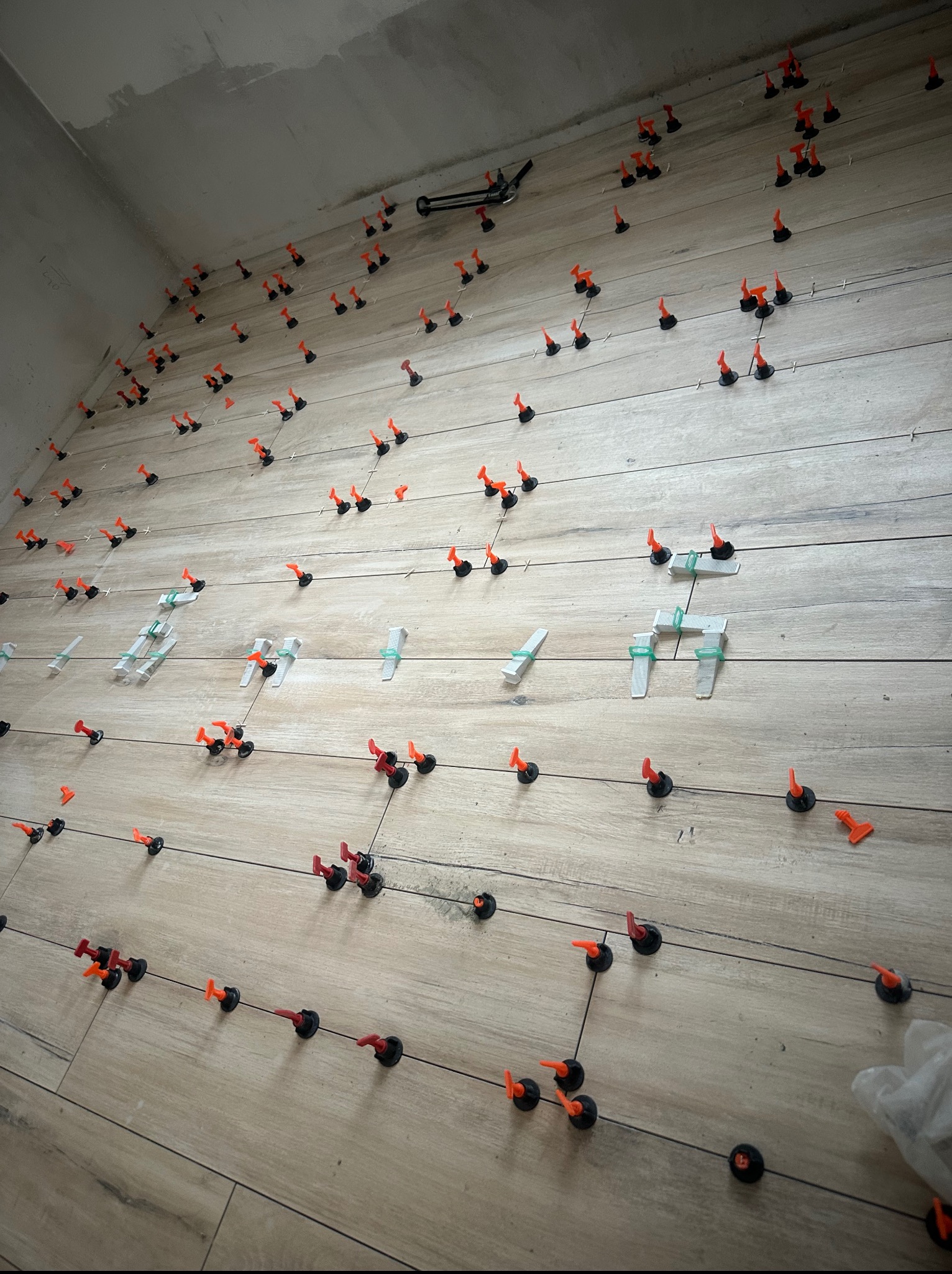 Układanie płytek podłogowych z systemem poziomowania, widoczne kliny i klipsy. Płytki imitujące drewno, jasne, ułożone poziomo. Ściana częściowo wykończona.