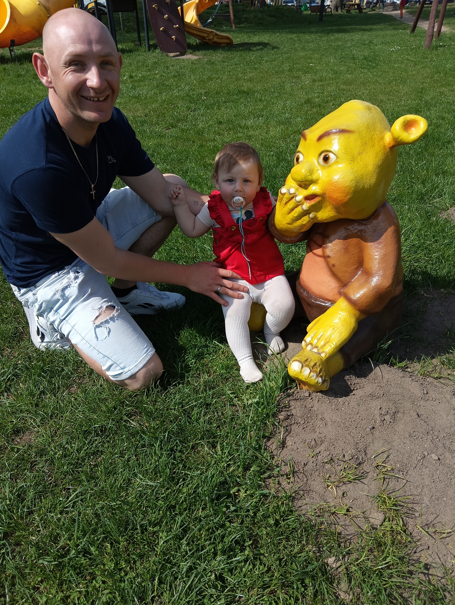 Uśmiechnięty mężczyzna w podartych jeansach i mała dziewczynka z smoczkiem pozują obok pomalowanej figury Shreka na trawniku placu zabaw.