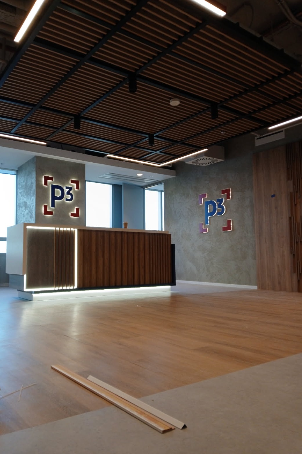 Nowoczesne wykończenie biura: drewniana lamelowa podłoga, recepcja z podświetleniem LED i logo firmy na ścianie. Widoczne listwy przypodłogowe.