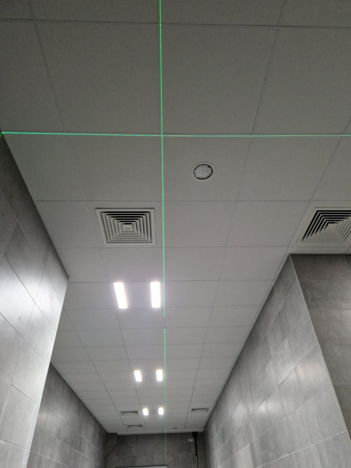 Sufit podwieszany z płyt w biurze, oświetlenie LED, kratki wentylacyjne i zielone linie lasera wyznaczające poziomy podczas montażu. Szare płytki na ścianach.