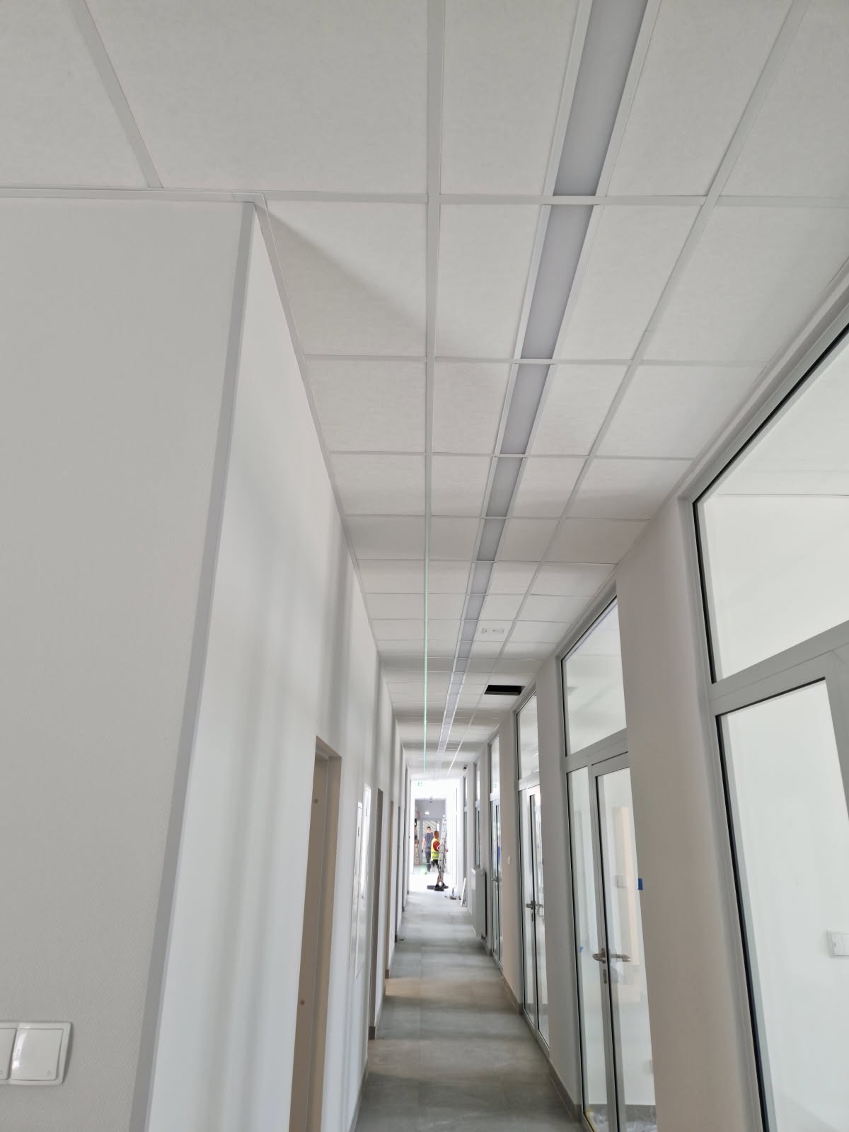Korytarz biurowy z podwieszanym sufitem kasetonowym i oświetleniem liniowym. Widok na wprost, ściany białe, szara podłoga, przeszklone drzwi po prawej.