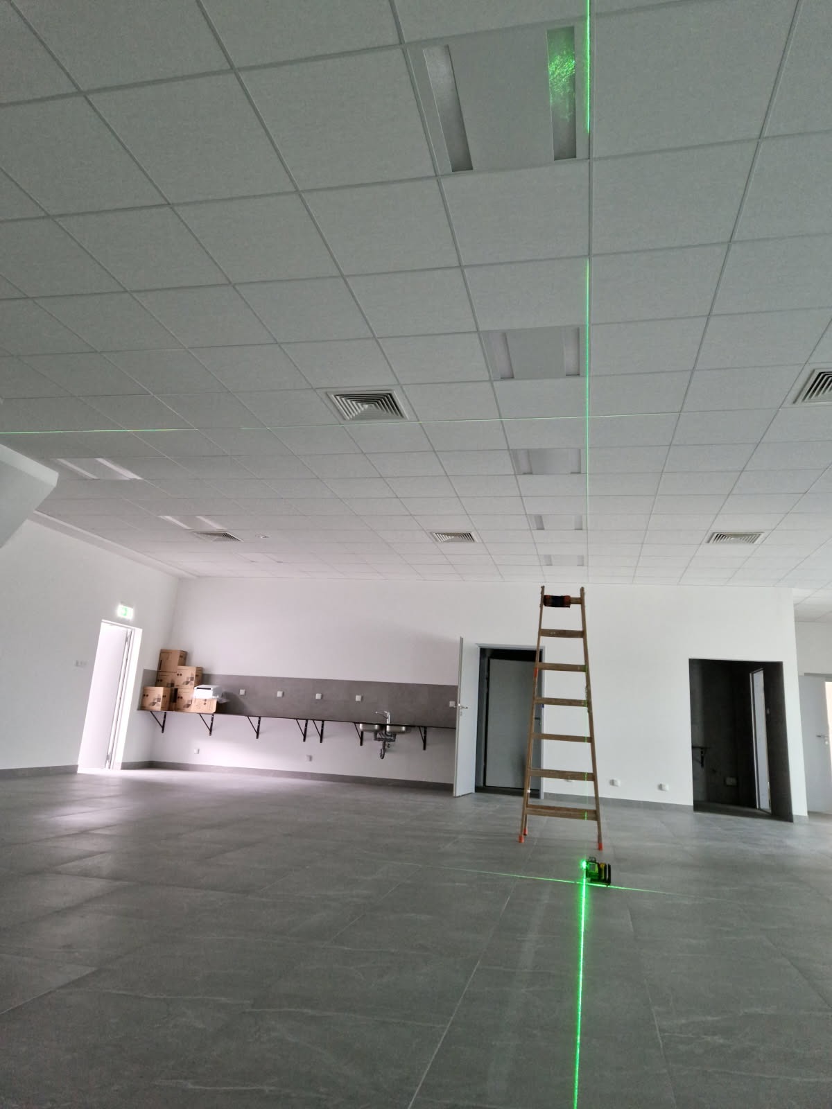 Duża sala z podwieszanym sufitem w trakcie montażu. Widoczna drabina i laser krzyżowy wyznaczający poziom. Szare płytki na podłodze i białe ściany.