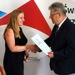 KANCELARIA ADWOKACKA ADWOKAT JUSTYNA MUSIELSKA - Uśmiechnięta kobieta w czarnej sukience wita się z mężczyzną w garniturze, wręczającym jej dokument na tle logo Urzędu Wojewódzkiego w Kielcach.