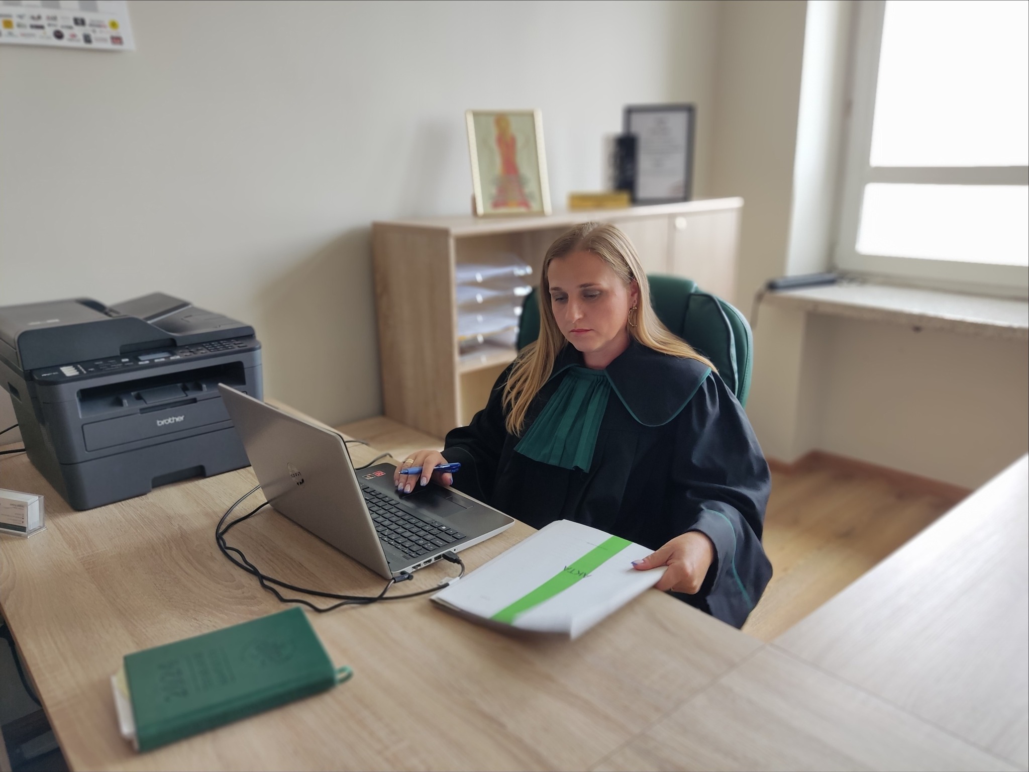 Adwokatka w todze pracuje przy biurku z laptopem, analizując akta sprawy. W tle biuro z drukarką i regałem.