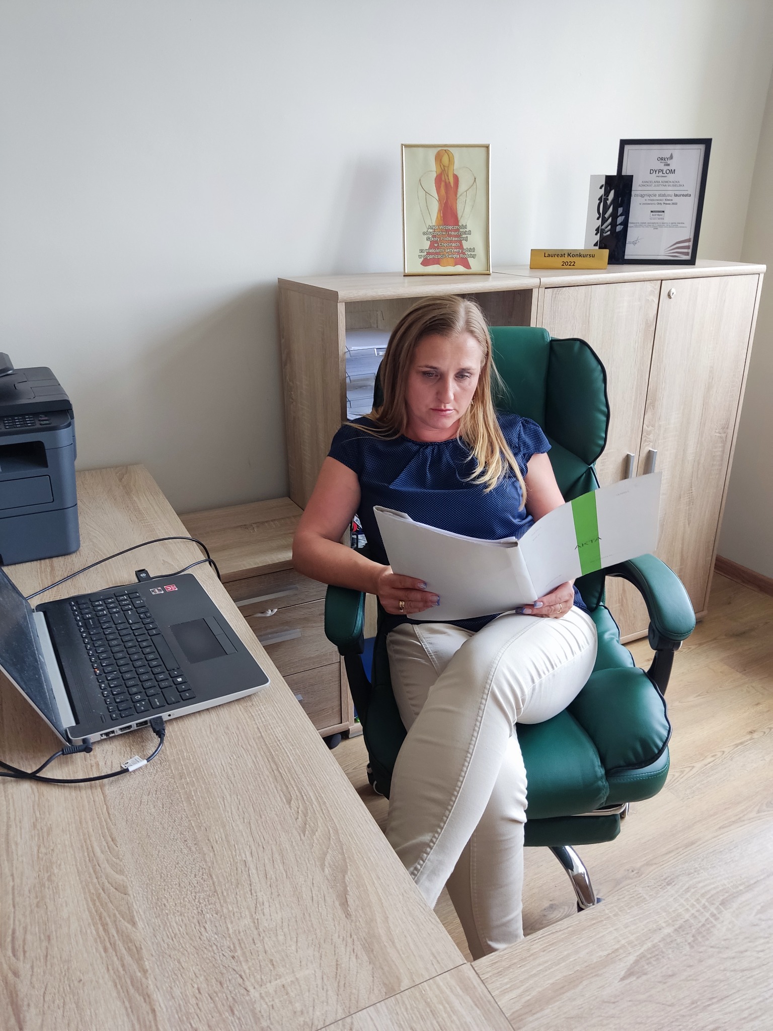 Adwokatka w biurze, siedzi na zielonym fotelu i analizuje akta. Na biurku laptop, w tle półka z dyplomem i nagrodą 'Laureat Konkursu 2022'.