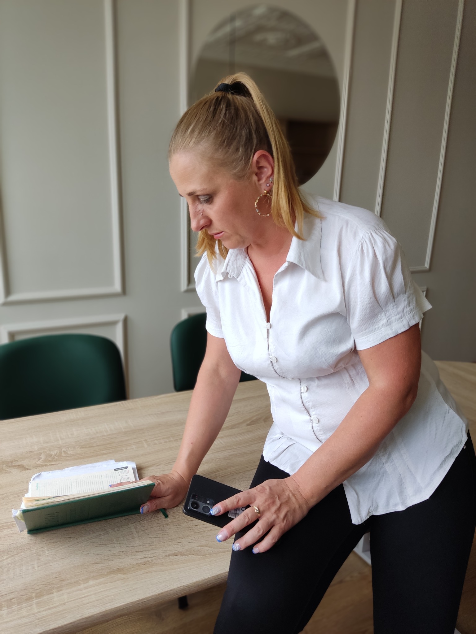 Kobieta w białej koszuli przegląda notatki i telefon na jasnym stole w biurze. W tle widoczne krzesła i dekoracyjna ściana.