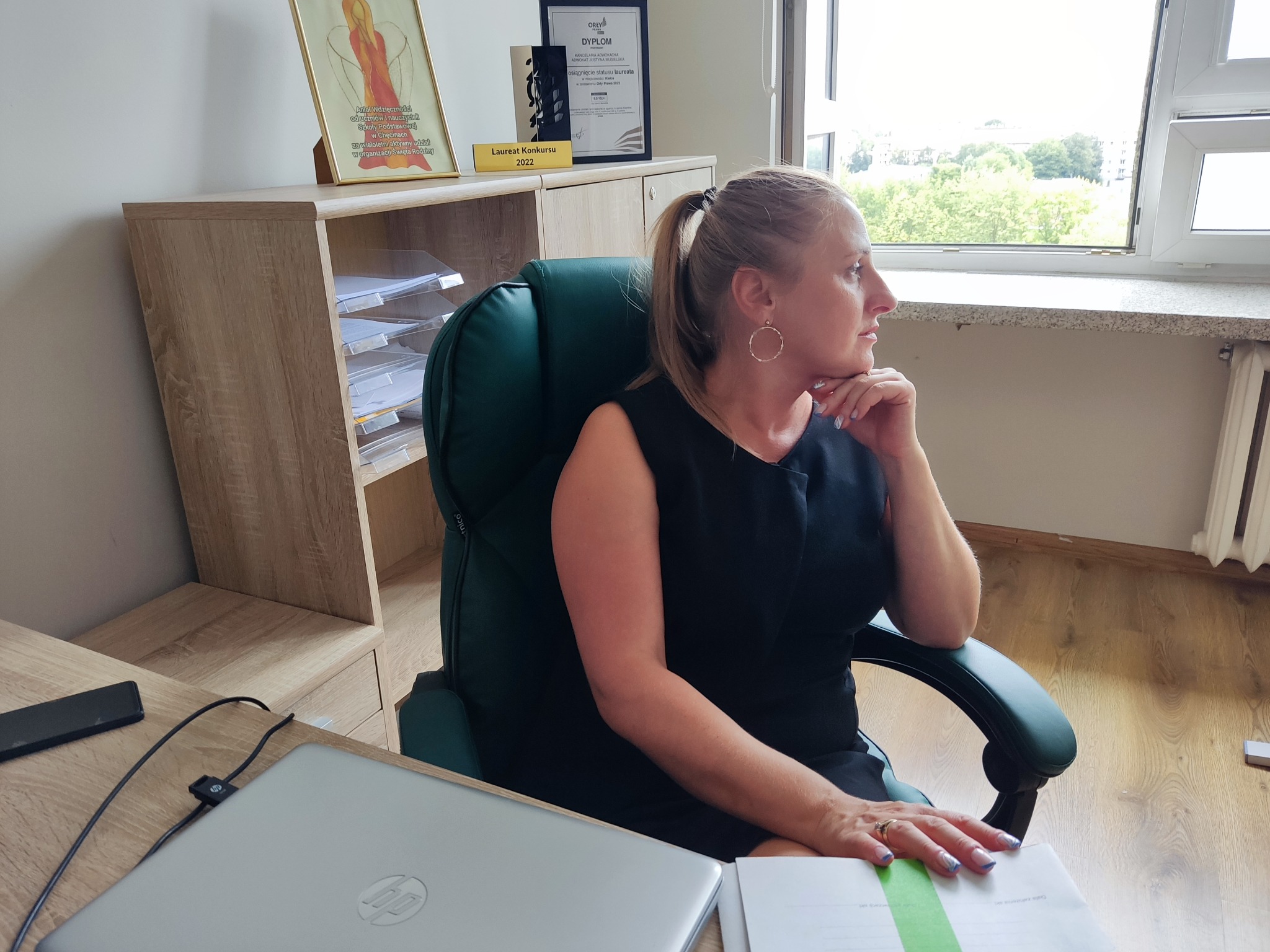 Adwokatka Justyna Musielska w biurze: dyplom Orły Prawa 2022, laptop HP, widok z okna. Kobieta w czarnej sukience zamyślona patrzy w dal.