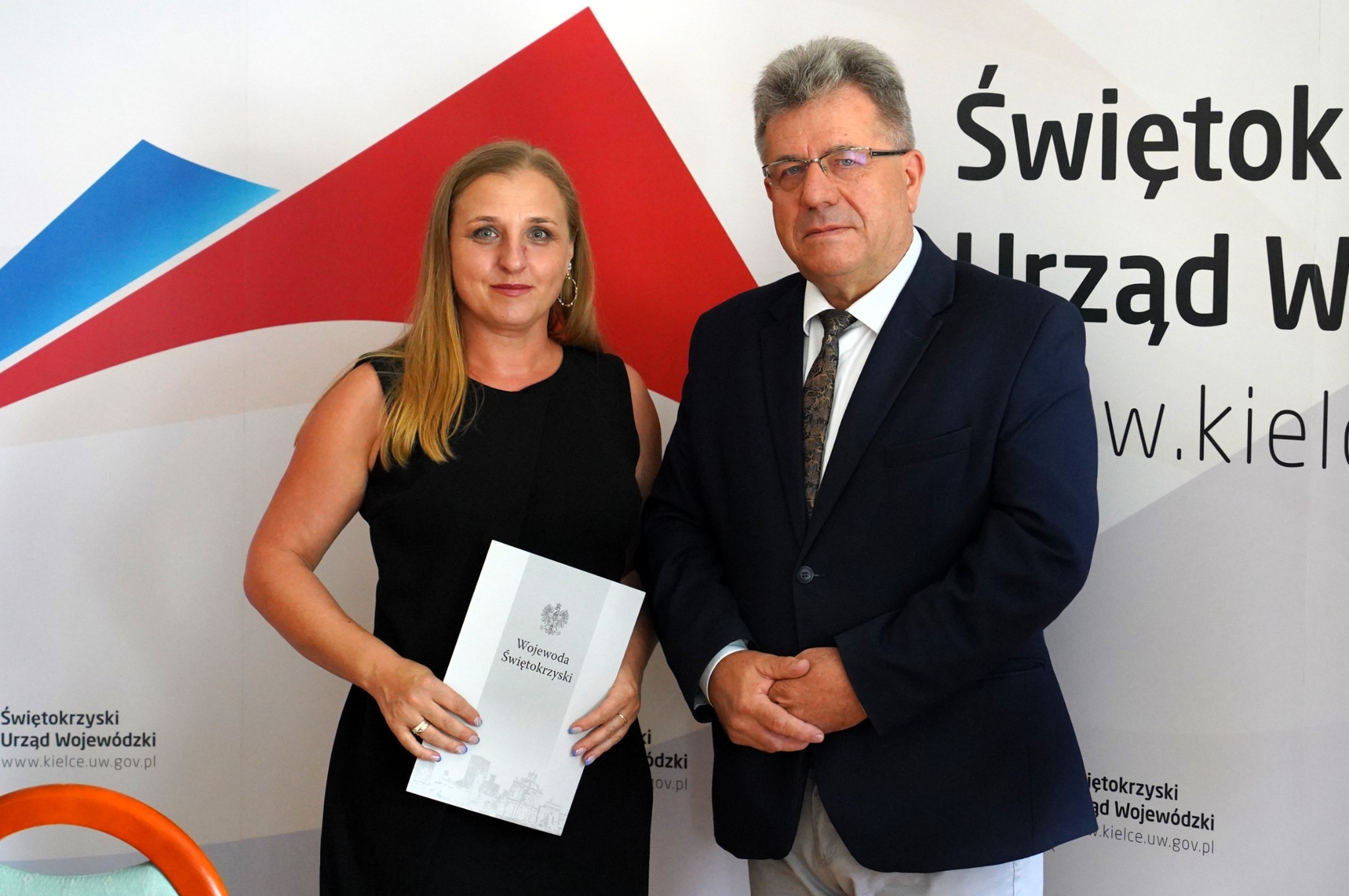 Dwie osoby, kobieta trzyma dokument Wojewody Świętokrzyskiego, mężczyzna w garniturze, na tle logotypu Urzędu Wojewódzkiego w Kielcach.