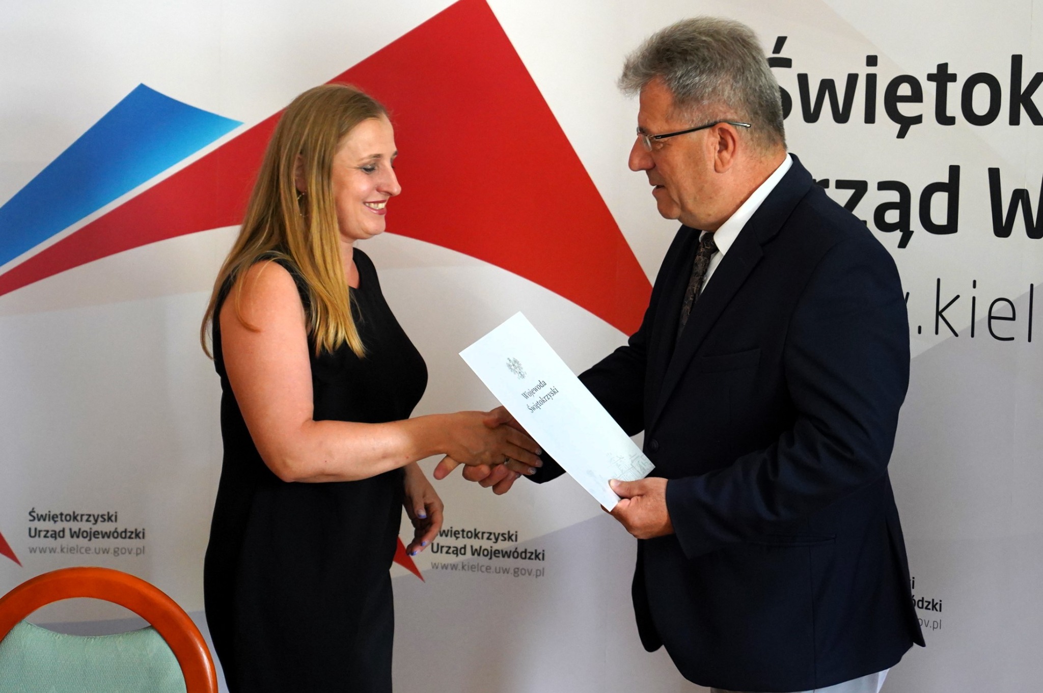 Uśmiechnięta kobieta w czarnej sukience wita się z mężczyzną w garniturze, wręczającym jej dokument na tle logo Urzędu Wojewódzkiego w Kielcach.