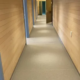 Długi korytarz wykończony drewnem z jasną, jednolitą posadzką. Widoczne drzwi z szarymi ościeżnicami i drewniany sufit z listew. Minimalistyczny design.