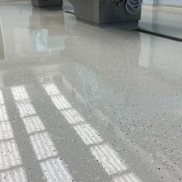 ProFloors - Błyszcząca, jasnoszara posadzka żywiczna z czarnymi i białymi drobinkami, odbijająca światło okien. Widoczny odpływ liniowy i fragmenty urządzeń.