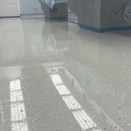 ProFloors - Błyszcząca, jasnoszara posadzka żywiczna z ciemnymi drobinkami w pomieszczeniu o białych kafelkach i sprzęcie medycznym. Widoczne odbicie światła w posadzce.