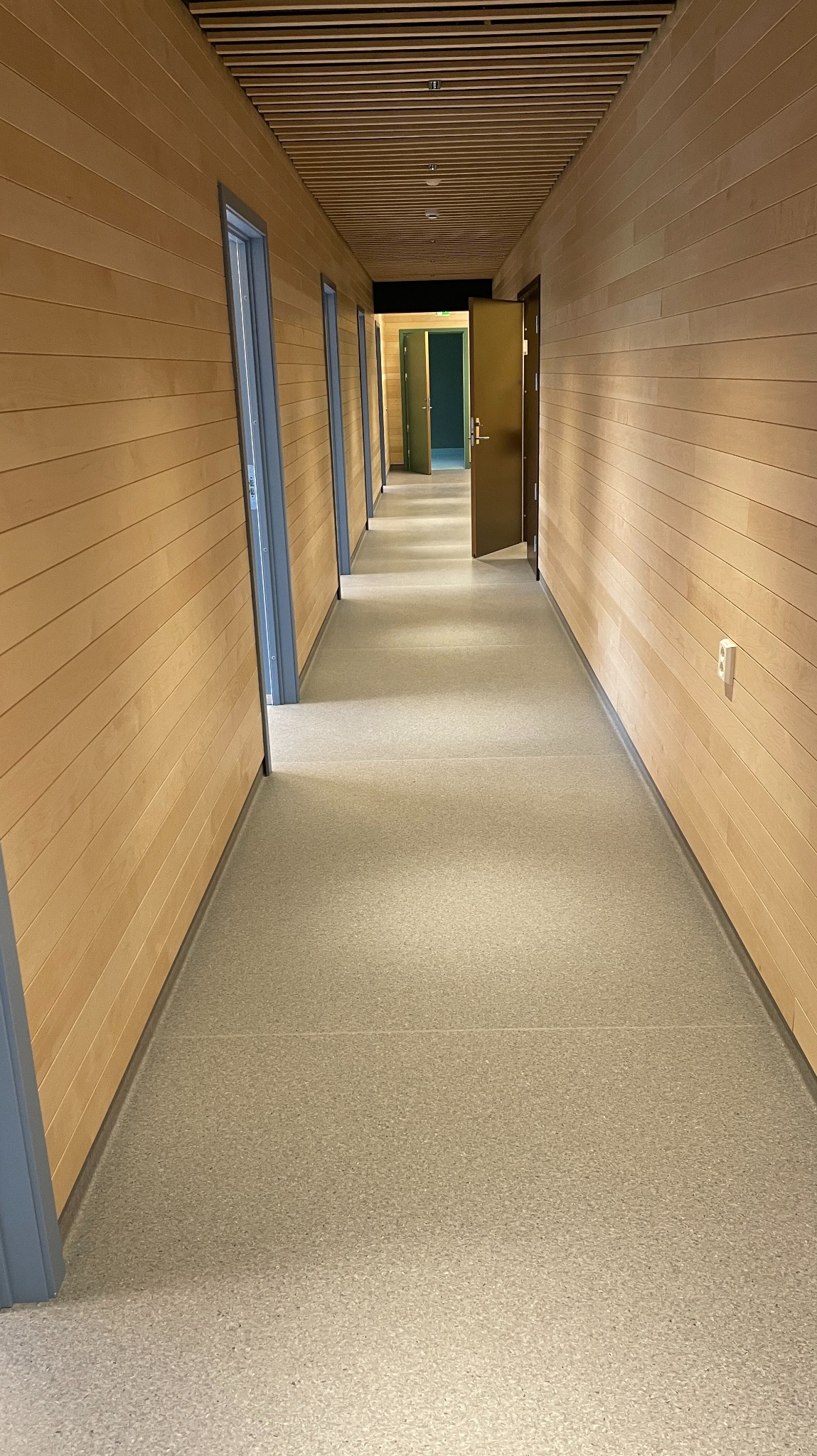 Długi korytarz wykończony drewnem z jasną, jednolitą posadzką. Widoczne drzwi z szarymi ościeżnicami i drewniany sufit z listew. Minimalistyczny design.