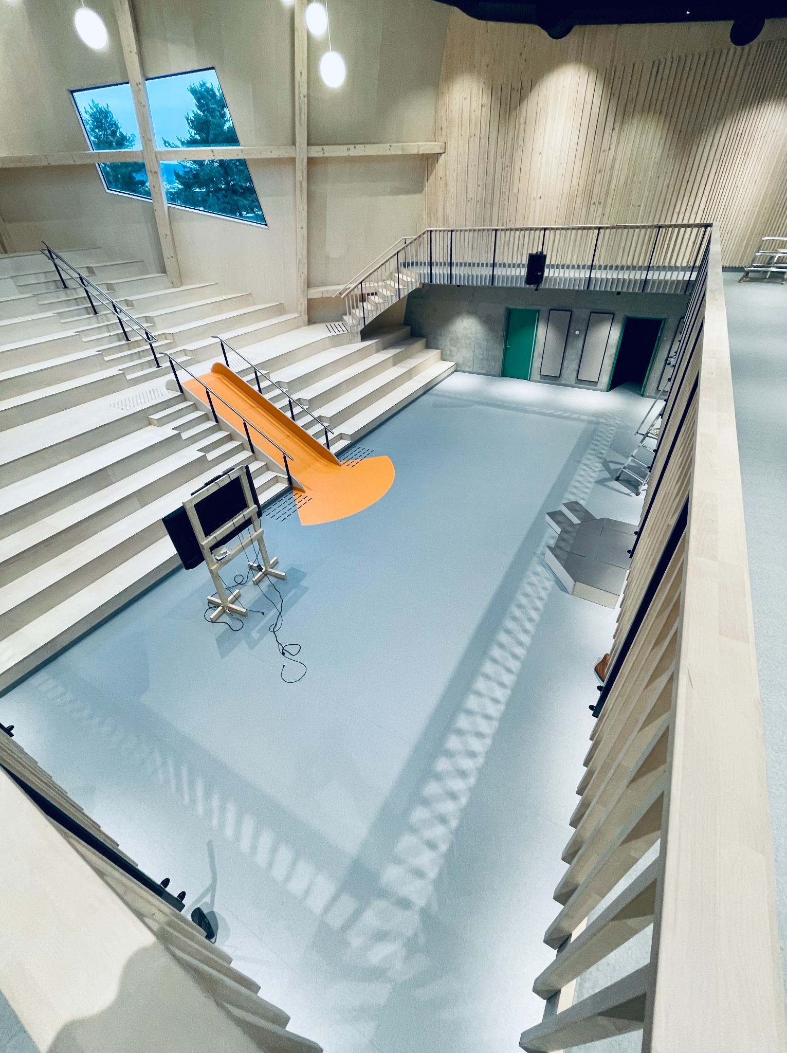 Jasna, nowoczesna przestrzeń z błękitną posadzką, schodami amfiteatralnymi i drewnianymi elementami. Oryginalne okno z widokiem na drzewa. Nietypowy, pomarańczowy element dekoracyjny.