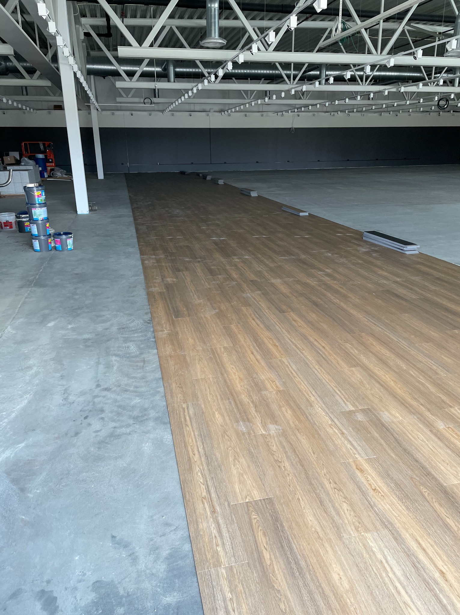 Wnętrze hali w trakcie układania paneli podłogowych imitujących drewno. Część podłogi surowa, betonowa. Na ścianie szara farba. Widoczne puszki z farbą.
