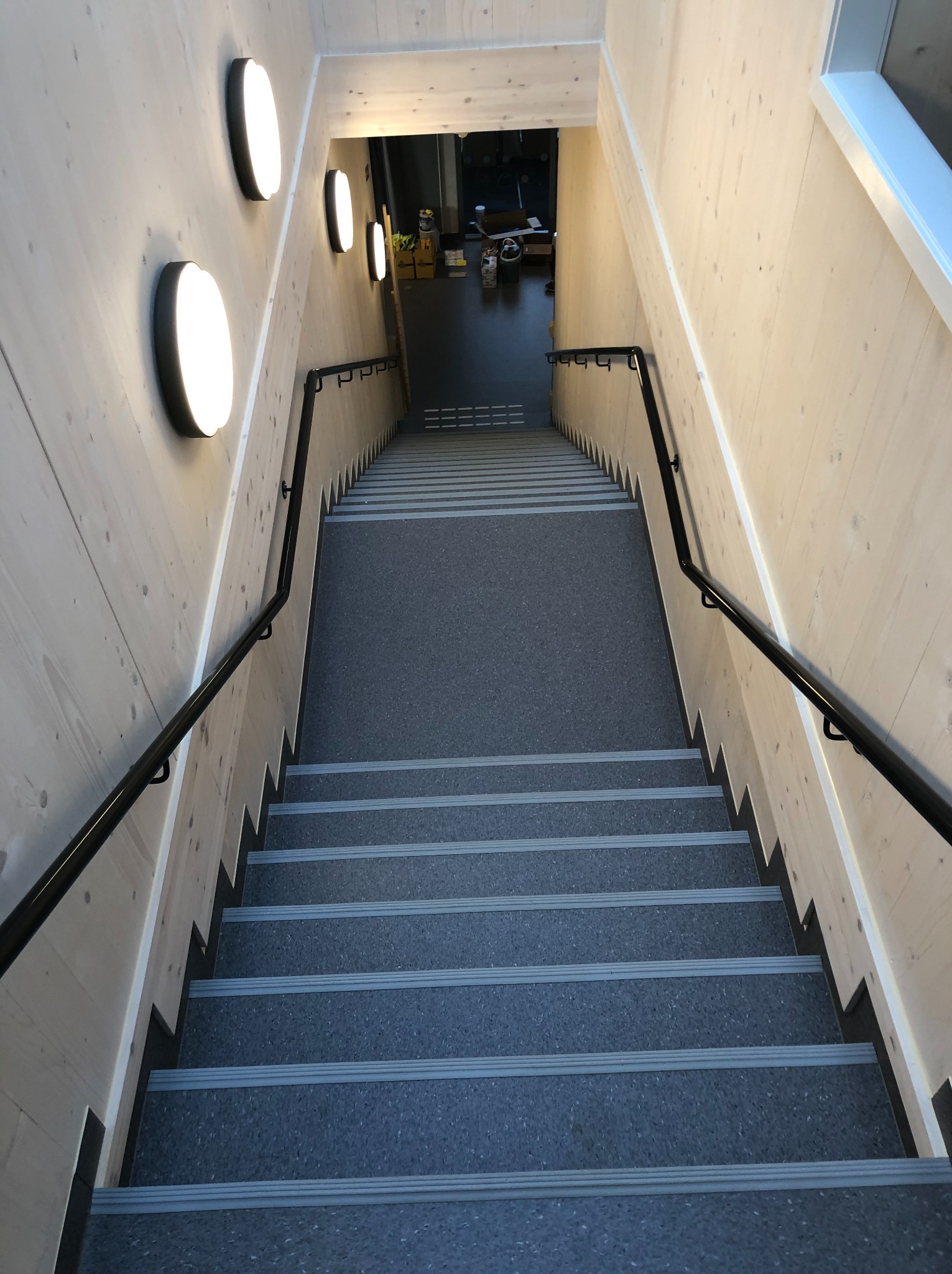 Schody wyłożone szarą wykładziną z antypoślizgowymi paskami, oświetlone okrągłymi lampami na drewnianej ścianie. Widok z góry na dół.