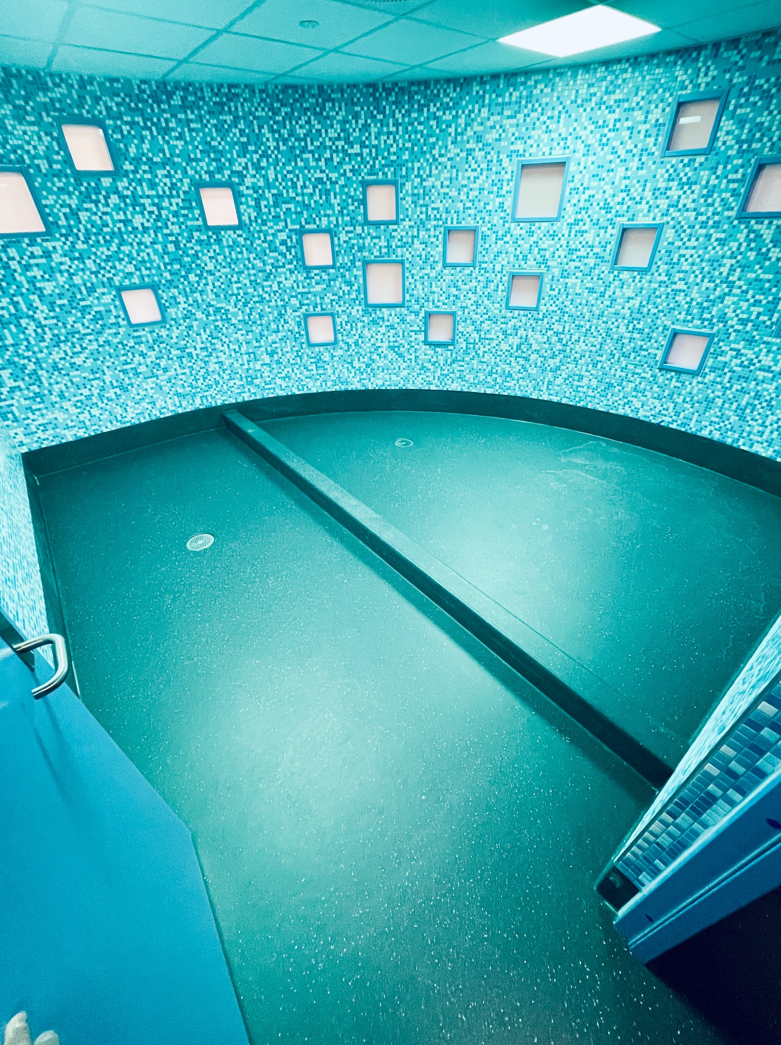 Turkusowa łazienka wykończona mozaiką i gładką posadzką, z oświetleniem w ramkach na ścianach. Widoczny odpływ liniowy i fragment schodów.