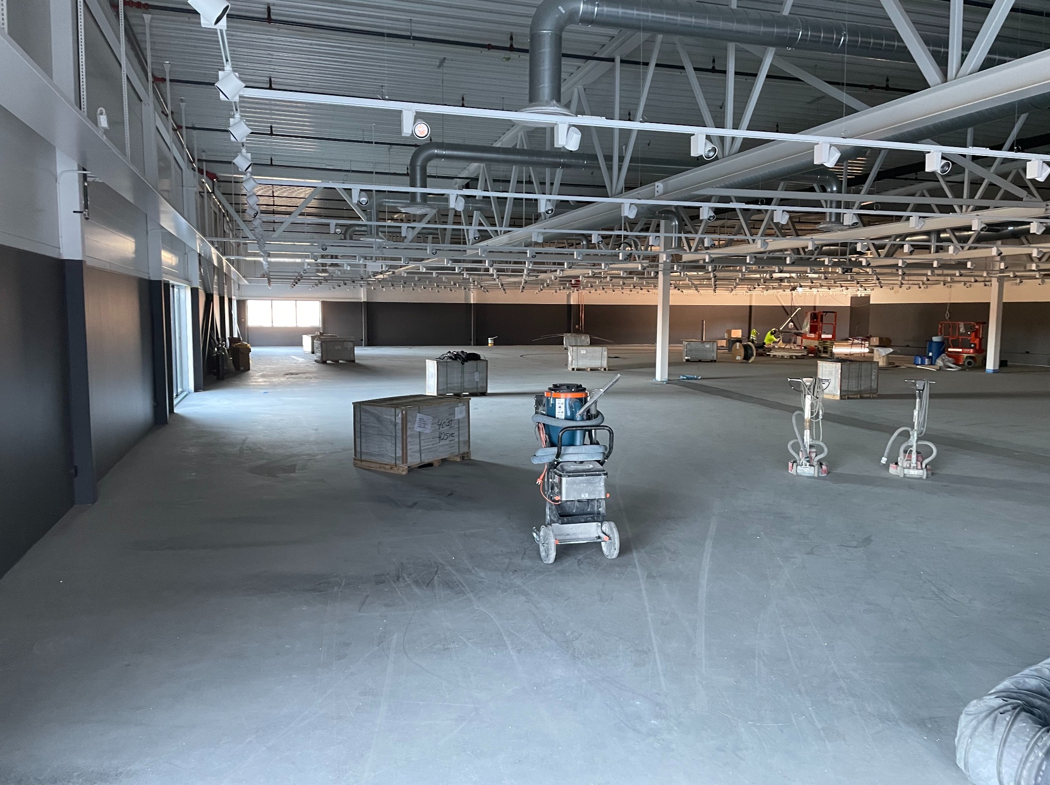 Duża hala z szarą posadzką. Widoczne maszyny do obróbki posadzek, palety z materiałami, pracownicy w oddali. Oświetlenie sufitowe równomiernie rozproszone.