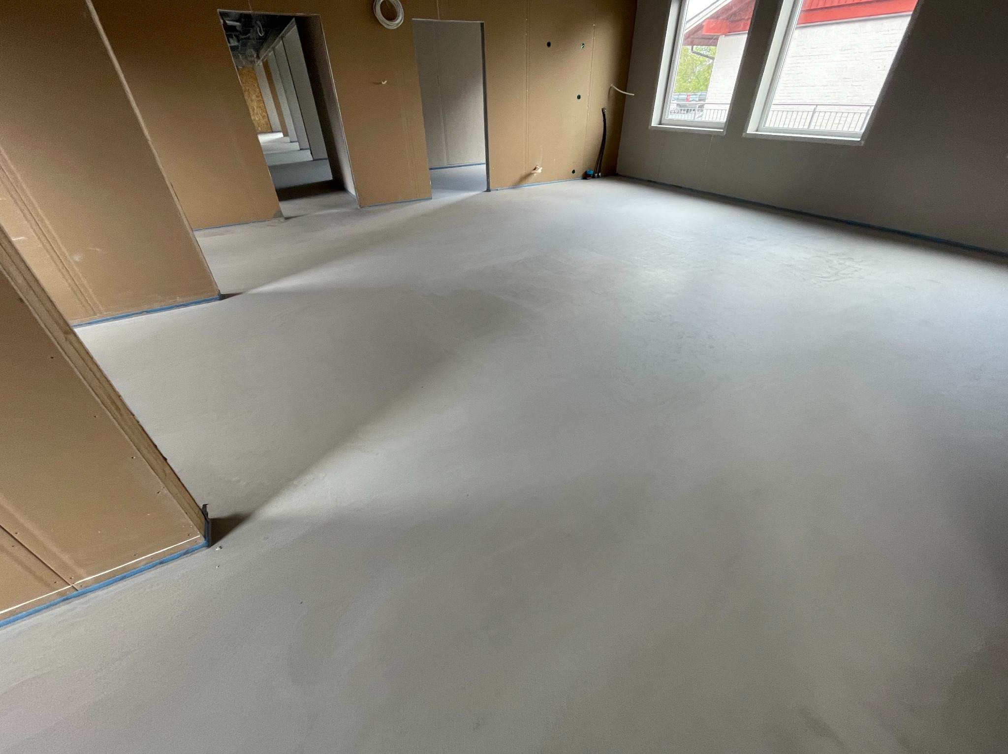 Wnętrze w trakcie prac wykończeniowych: świeżo wylana, gładka, jasna posadzka z widocznymi ścianami w surowym stanie i taśmą zabezpieczającą. W tle widoczne okna.