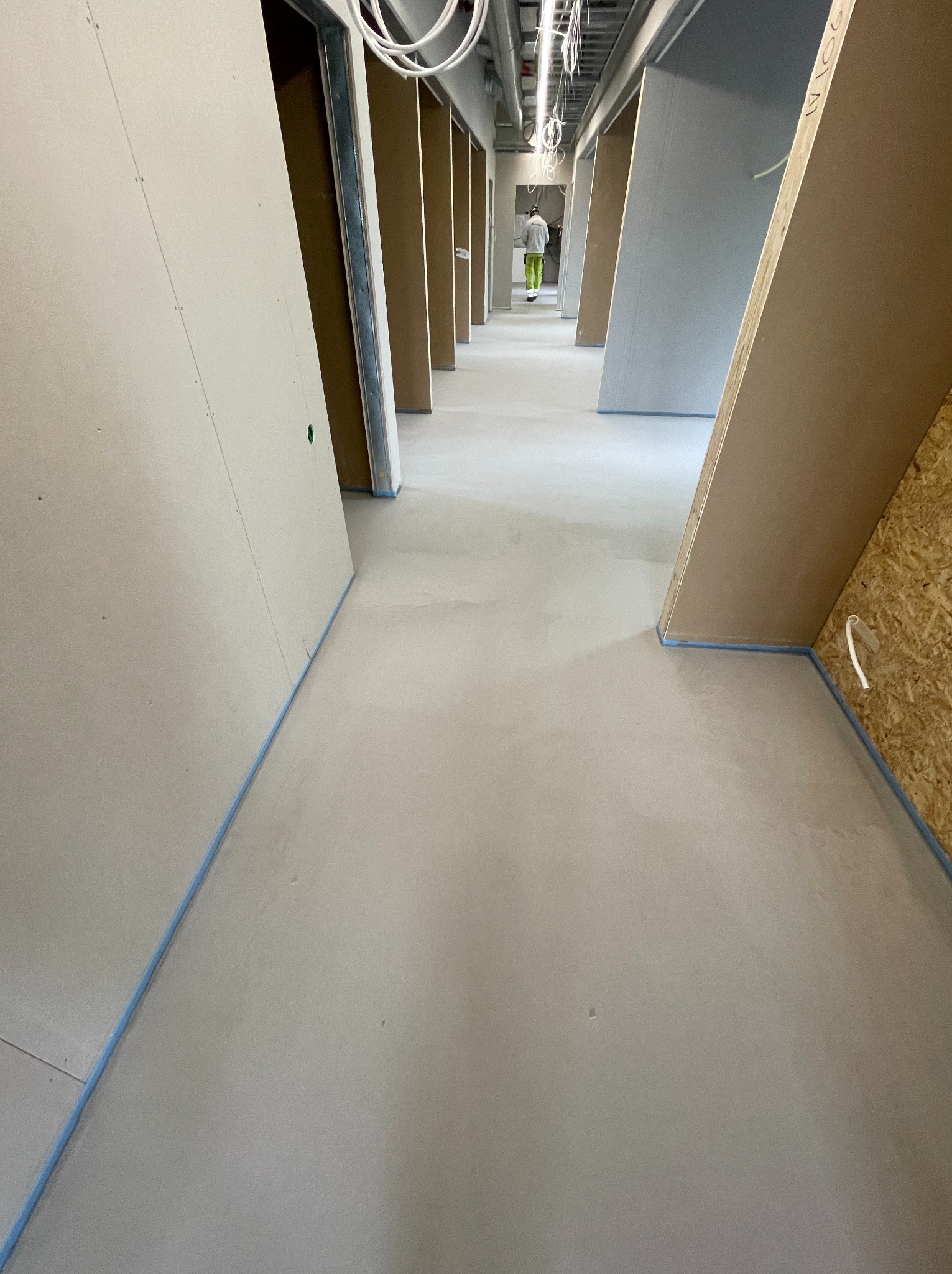 Szara, gładka posadzka w długim korytarzu z surowymi ścianami i widoczną instalacją pod sufitem. W oddali postać w odblaskowej kamizelce.