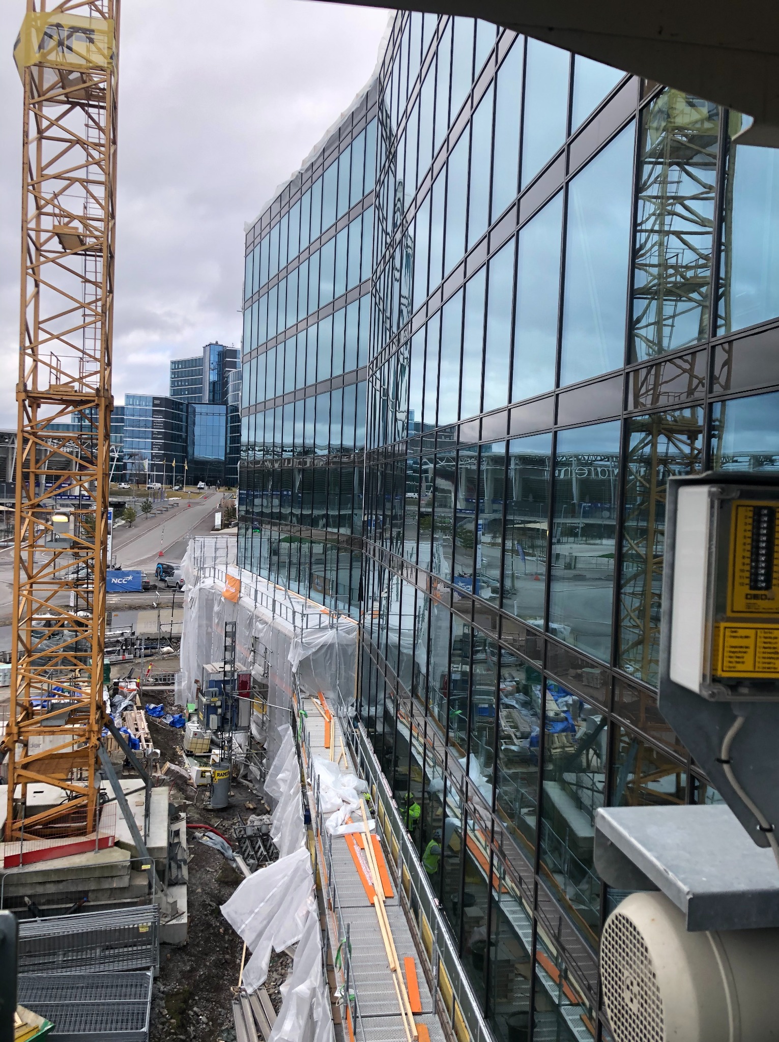 Budynek biurowy w trakcie budowy z widocznym żurawiem i rusztowaniami. Fasada szklana odbija otoczenie. Prace wykończeniowe w toku, widoczne materiały budowlane.