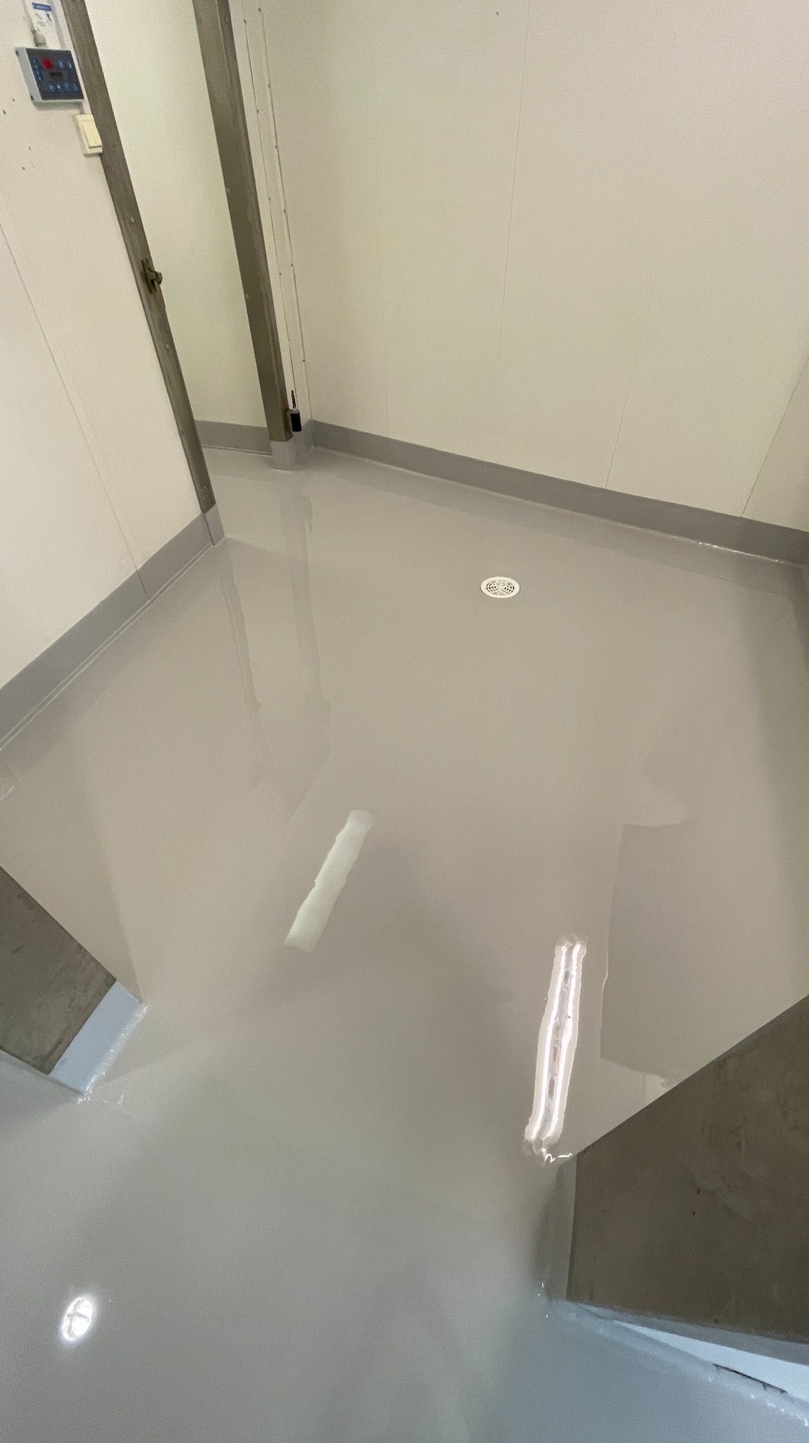 Jasnoszara, gładka posadzka żywiczna w pomieszczeniu z odpływem podłogowym i panelami ściennymi. Widoczne odbicia światła na powierzchni.