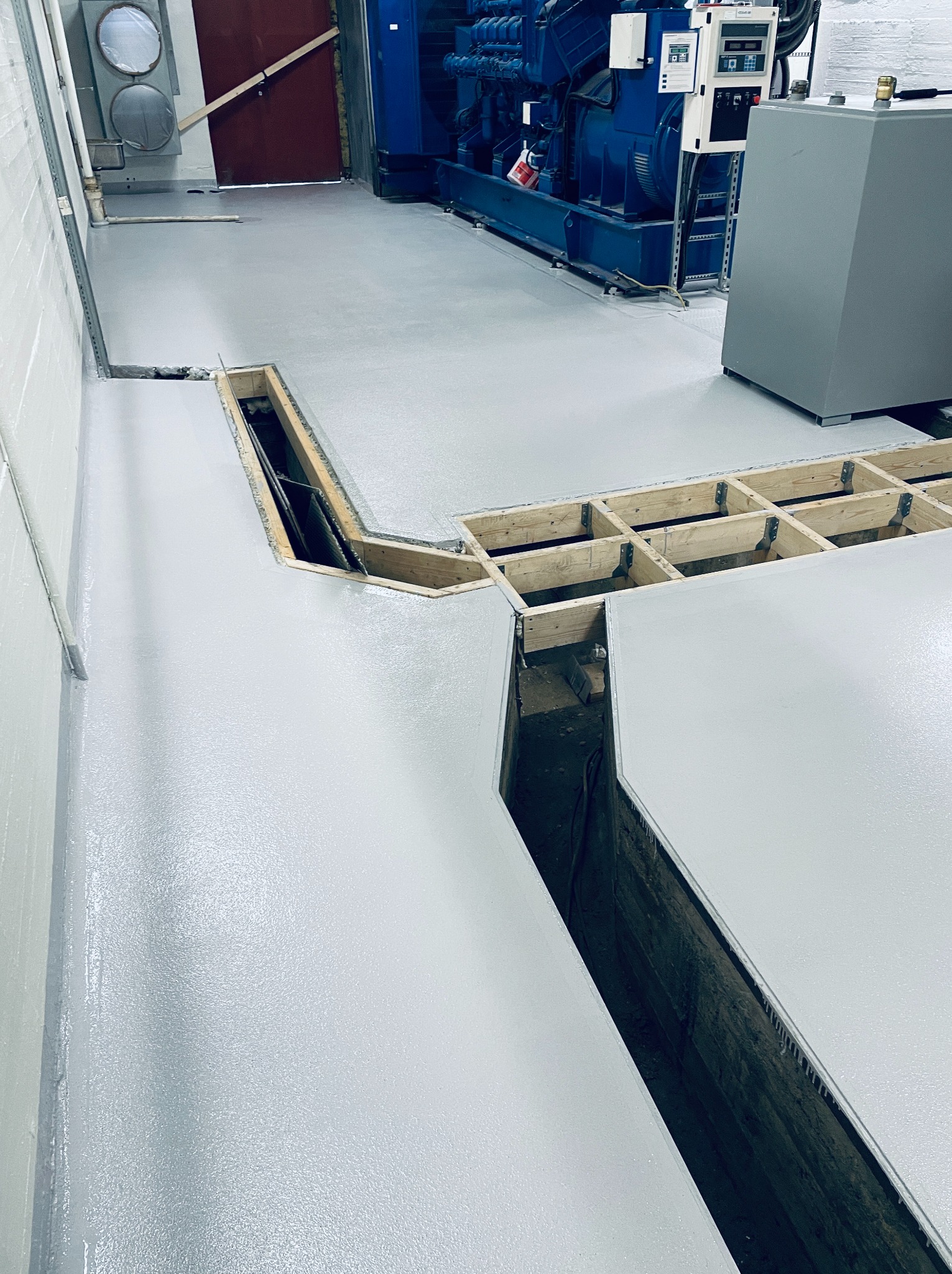 Jasnoszara, błyszcząca posadzka przemysłowa z widocznymi kanałami technicznymi i fragmentami drewnianej konstrukcji. W tle agregat prądotwórczy i instalacje.