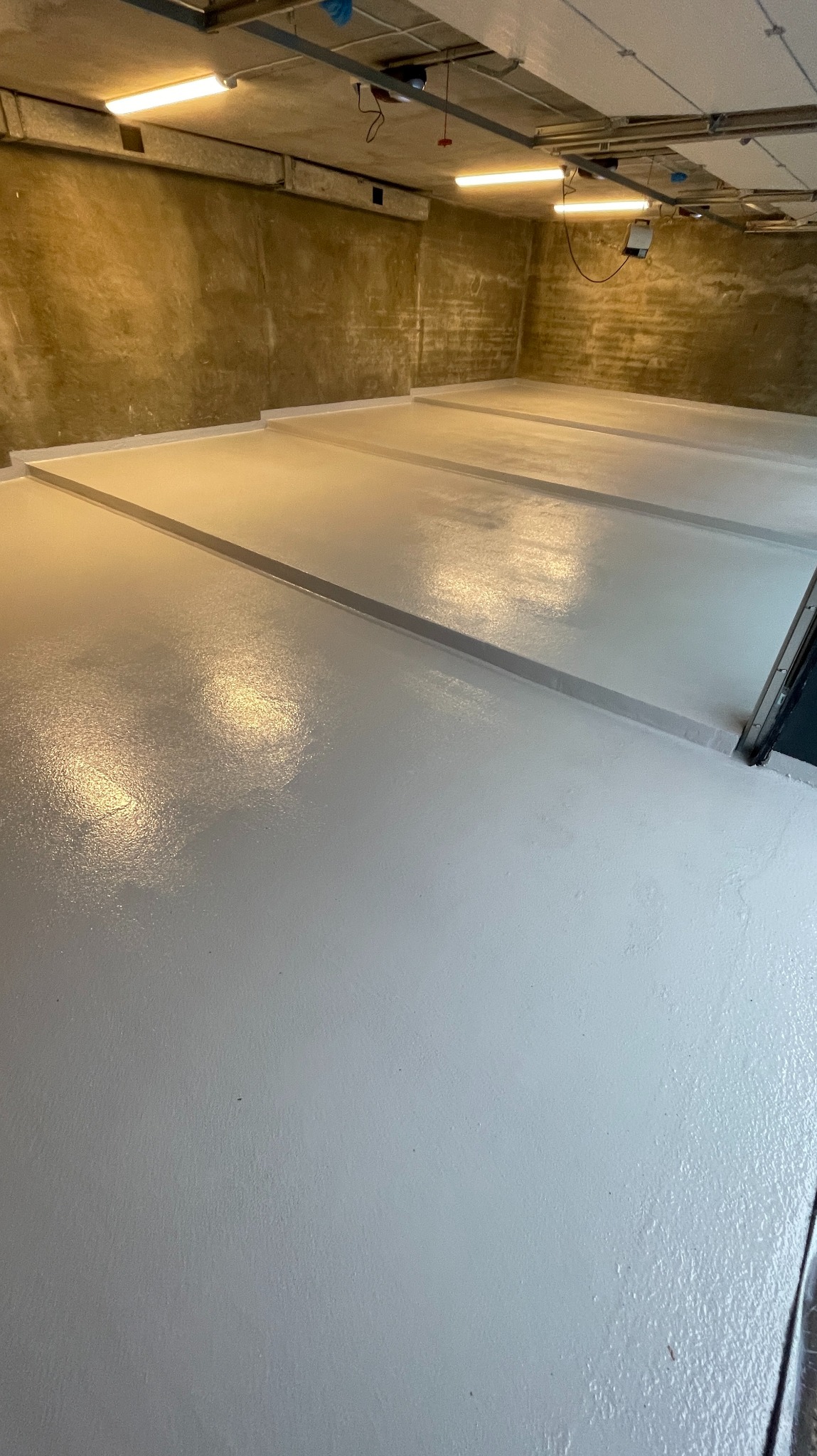 Jasna, świeżo wylana posadzka w garażu lub hali, z widocznymi dylatacjami. Surowe ściany w tle, oświetlenie jarzeniowe.